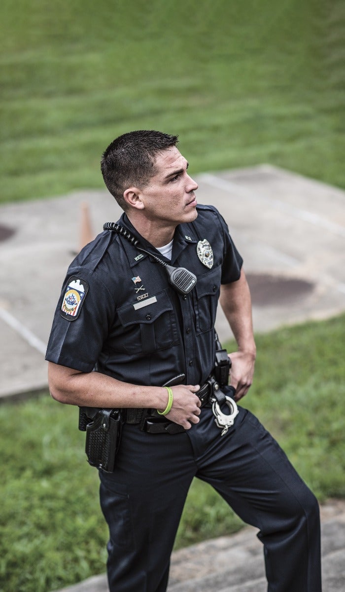 Photograph of a police officer standing outdoors near a grassy area with a lime green bracelet a black holster and visible handcuffs on his belt showcasing a detailed patch on his uniform