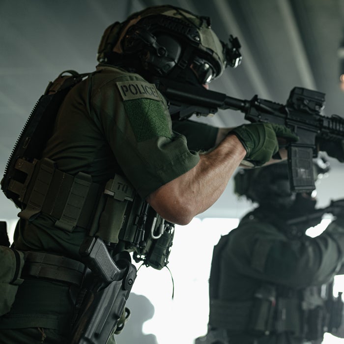 Photograph of a SWAT team member in action aiming an AR-15 style rifle indoors with a second officer in the background showcasing tactical gear, dark green uniforms, and a visible POLICE patch
