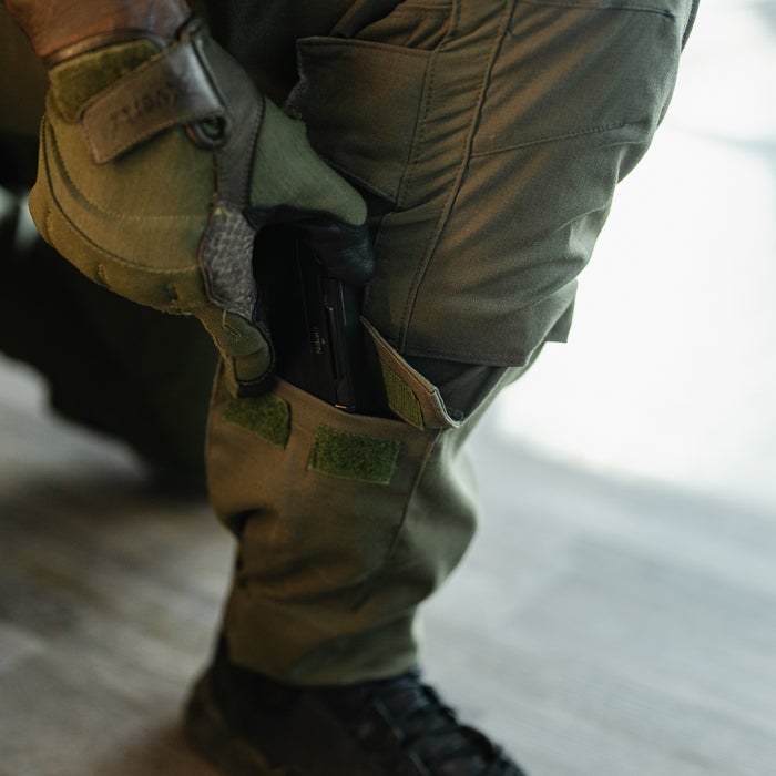 Photograph person inserting black handgun into olive green tactical pants leg featuring Velcro straps and dark gray tactical gloves.