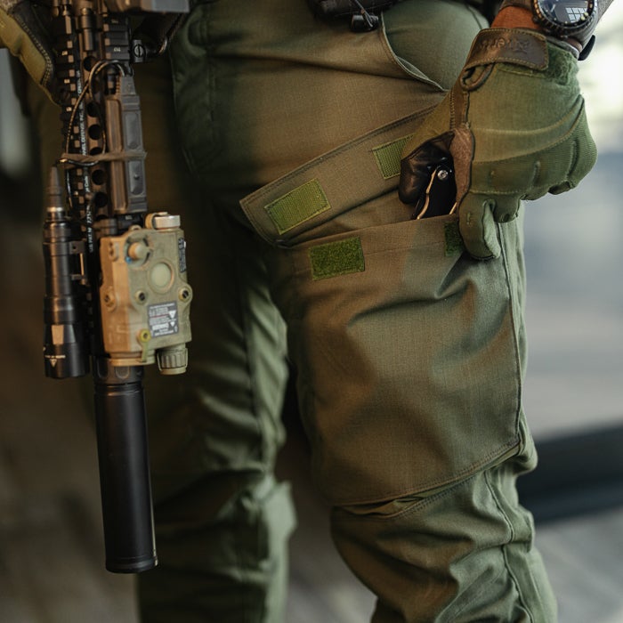 Photograph of olive green tactical pants with multiple pockets a flashlight and suppressor attached to a rifle showing velcro straps and a watch