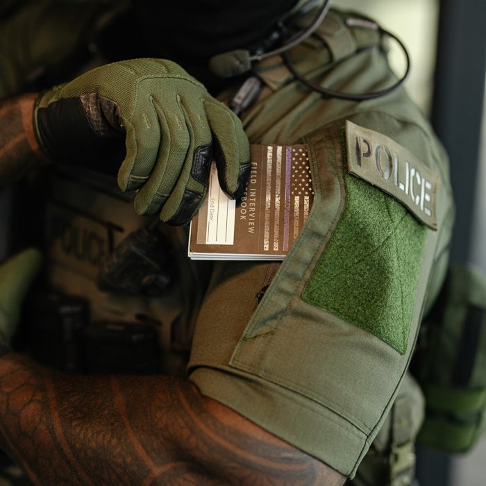 Photograph of a police officer's olive green uniform with a thin blue line field interview book in the pocket, showcasing tactical gloves and a detailed arm tattoo