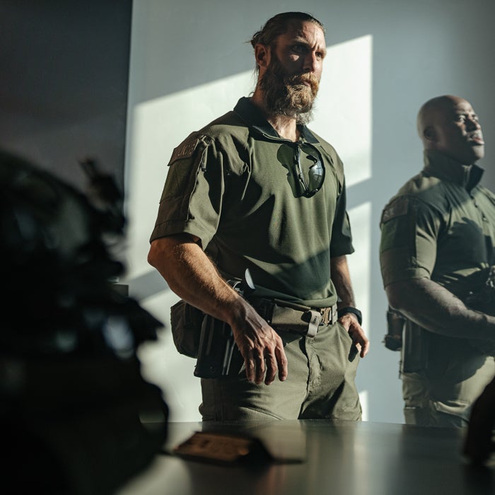 Photograph two SWAT officers in olive green tactical gear inside a building with dramatic sunlight, one with a prominent beard and sunglasses, showcasing their holstered weapons and serious expressions
