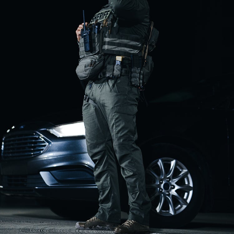 Photography: Sheriff standing beside a Ford Fusion at night wearing a tactical vest with a radio, multiple pouches, and a knife, showcasing his olive-green uniform and sturdy boots