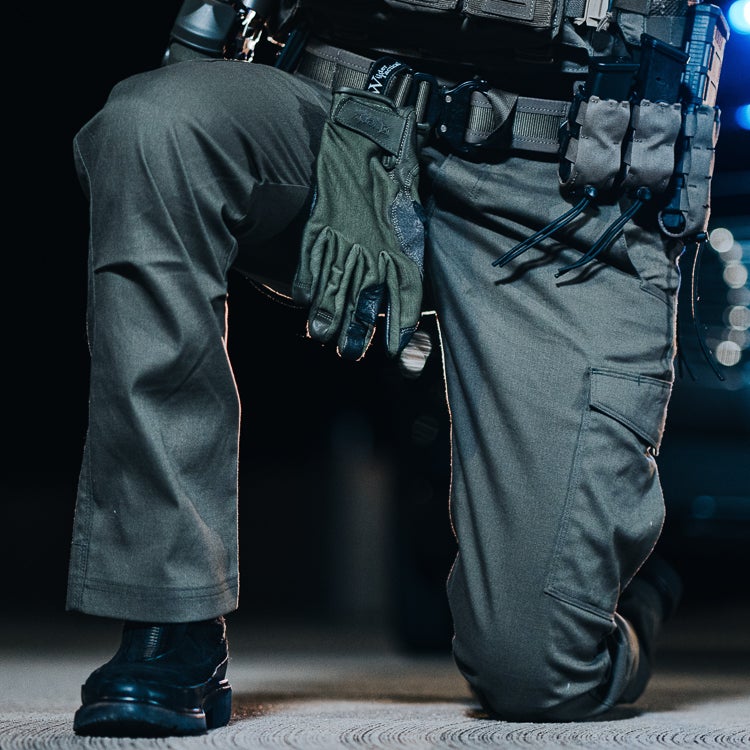 Photograph of a person kneeling outdoors wearing olive green tactical pants a black belt and a dark green tactical glove at night with blurry city lights in the background