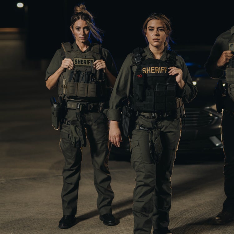 Photography two female sheriff's deputies in tactical gear standing at night near a police vehicle with visible "Sheriff" patches and utility pouches dark green uniforms and tactical belts