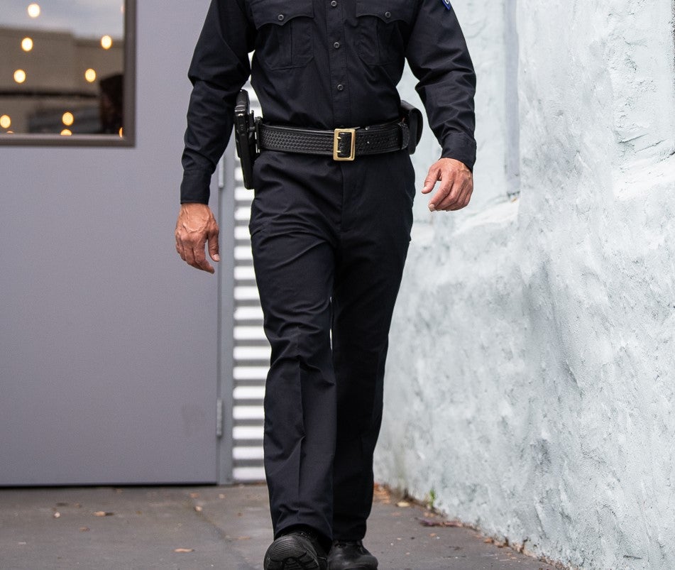 Photography Unknown Artist A uniformed security officer walks past a grey building with a glass door and string lights a gold belt buckle and black pants are visible