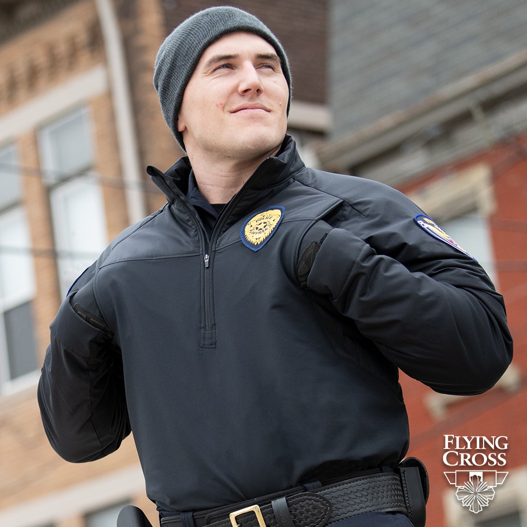 Photography Police officer in dark gray knit cap and black uniform shirt with gold badge stands outside a brick building with visible windows a partially visible brown brick building in the background