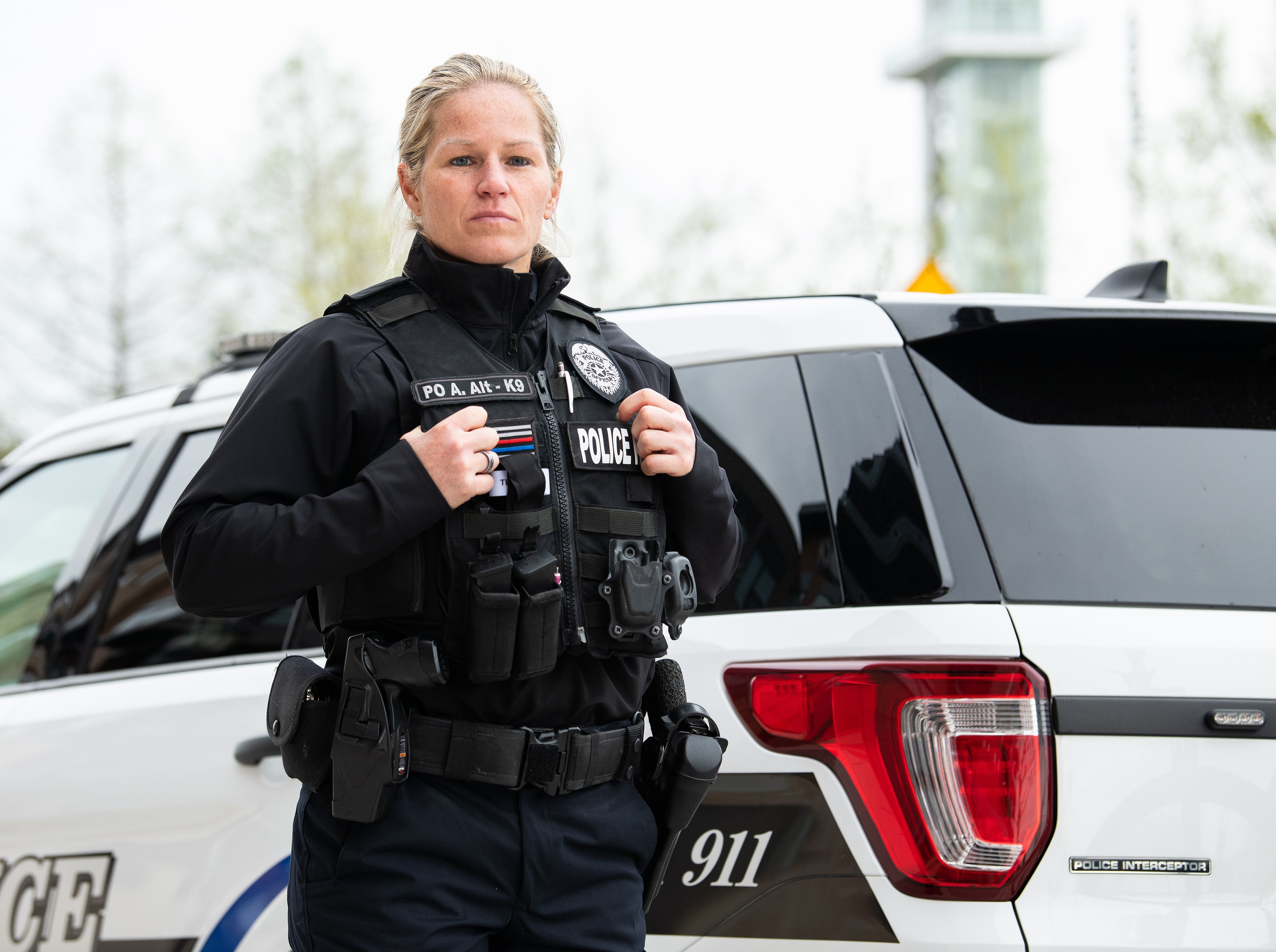 Photograph of a female police officer in uniform standing by a Police Interceptor SUV with a thin blue line patch, a police badge, and a PO Alt-K9 designation visible on her vest