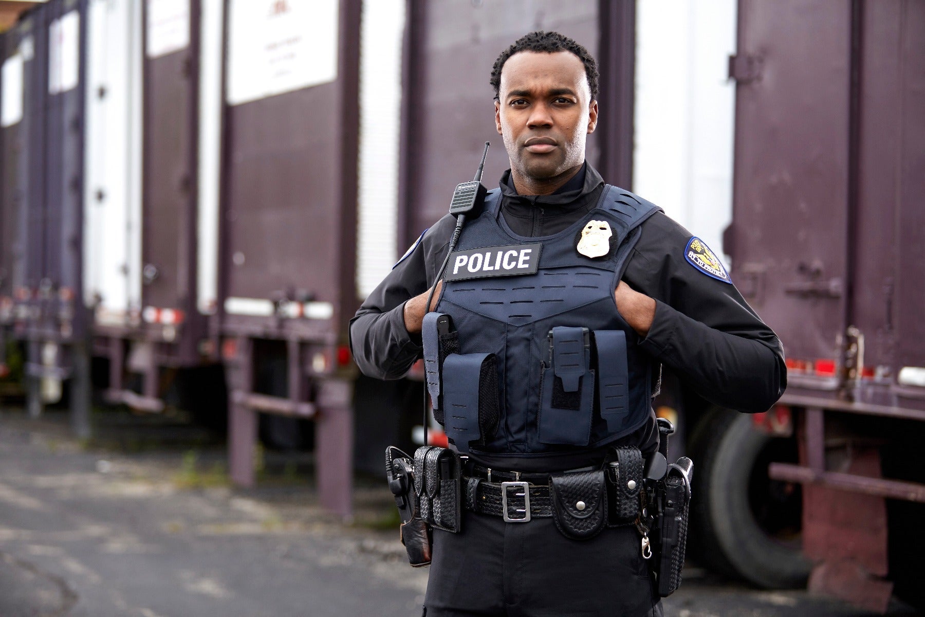 Photograph of a police officer in uniform standing near cargo containers with a police radio, a gold badge, and a dark blue tactical vest