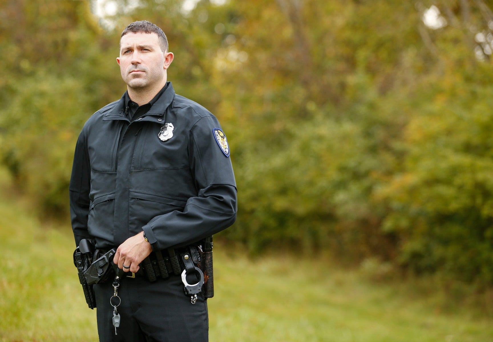 Photograph of a police officer in a black uniform outdoors with a gun and handcuffs, standing in a grassy field against a blurred background of autumnal trees with yellow and green foliage