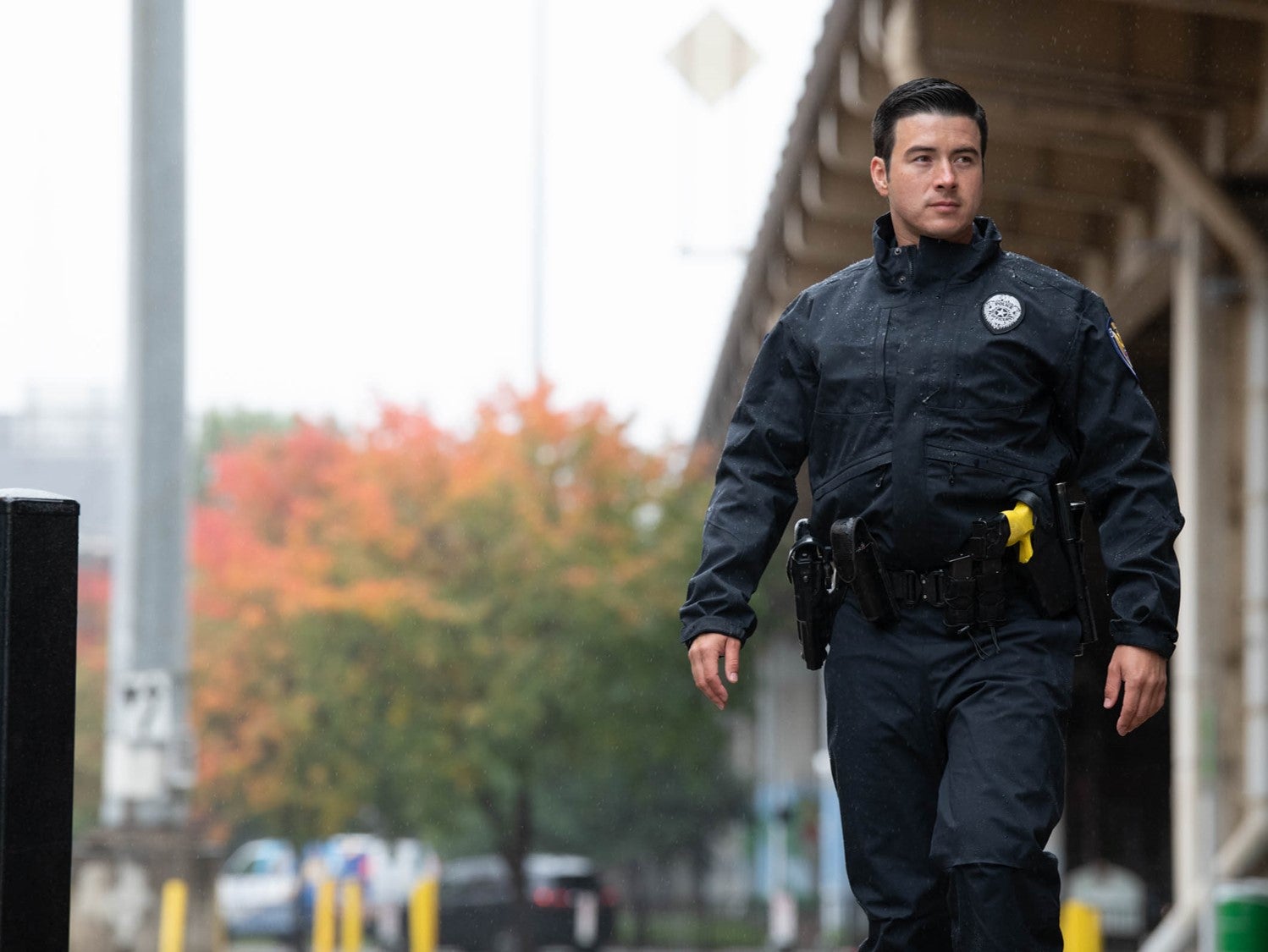 Photograph of a police officer walking in an urban setting featuring autumnal foliage vibrant yellow safety equipment and a raised overpass