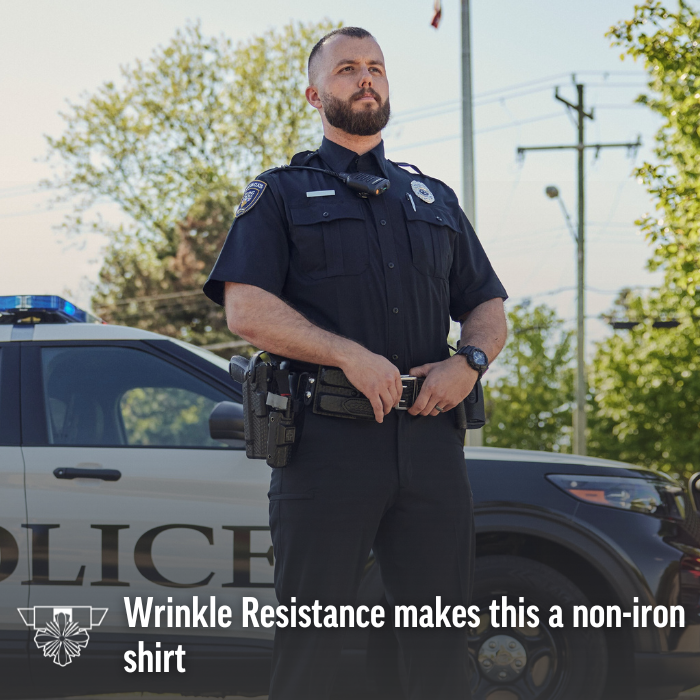 Photograph of a police officer in a wrinkle-resistant uniform standing by a police car near leafy trees and utility poles, showcasing his dark pants, black duty belt, and crisp shirt.
