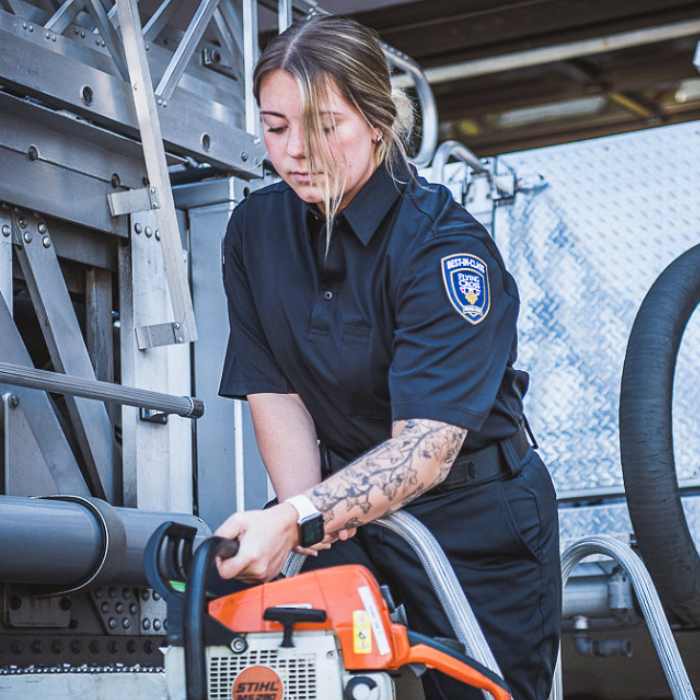 Photograph A female firefighter with a floral tattoo, wearing a black uniform with a Flying Cross Fire Department patch, prepares an orange Stihl chainsaw near a firetruck's ladder and hoses