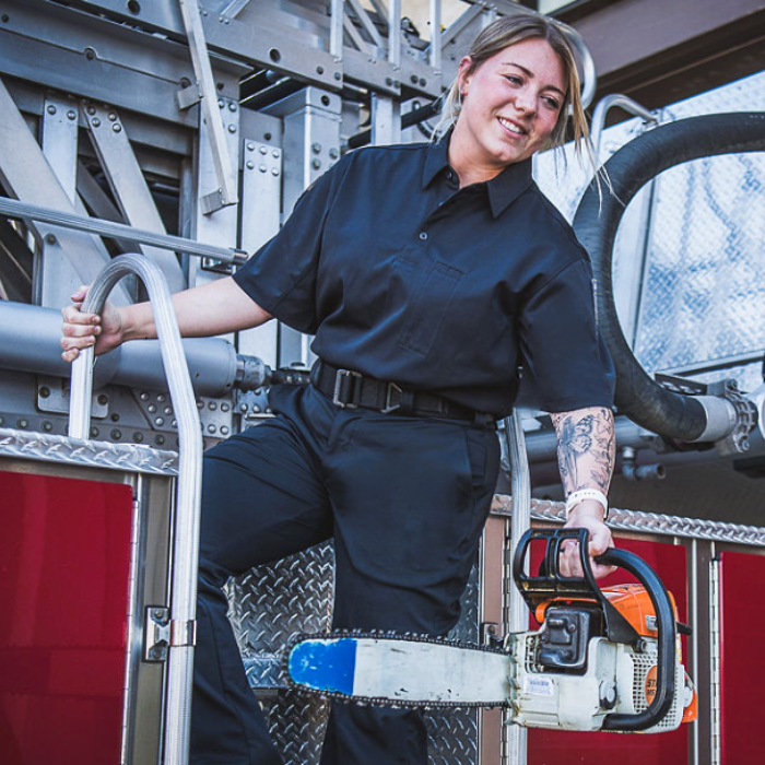 Photograph of a female firefighter on a firetruck ladder holding a chainsaw with a blue tip near red compartments and metallic grey components