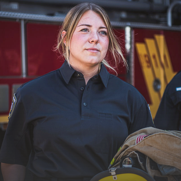 Photograph female firefighter in dark wrinkle-resistant polo shirt near firetruck with yellow stripes and red body holding folded fire hose and helmet showcasing embroidered badge and reflective details