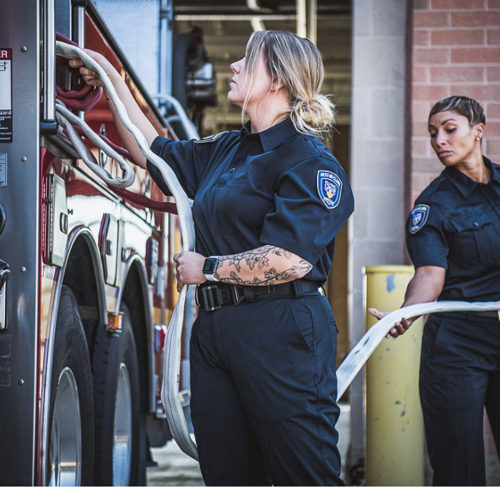 Photograph two firefighters at a fire station preparing a fire hose near a red firetruck with visible tires and a brick building showing a woman with a floral forearm tattoo and a smartwatch