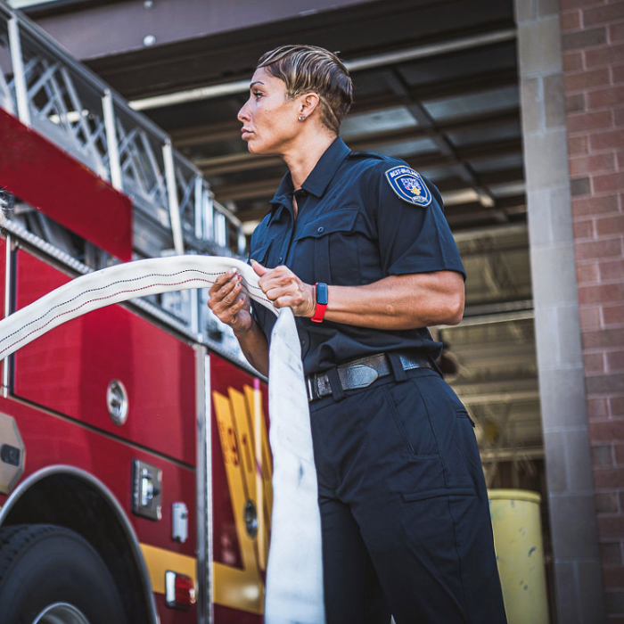 Photograph female firefighter holding a firehose in front of a red firetruck with yellow stripes and a West Hills Fire Department patch, showcasing her muscular arms, a red fitness tracker and a short, stylish haircut