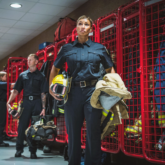 Photograph of a female firefighter in a fire station hallway holding a helmet and jacket amidst bright red lockers and fellow firefighters with yellow helmet and tan jacket details