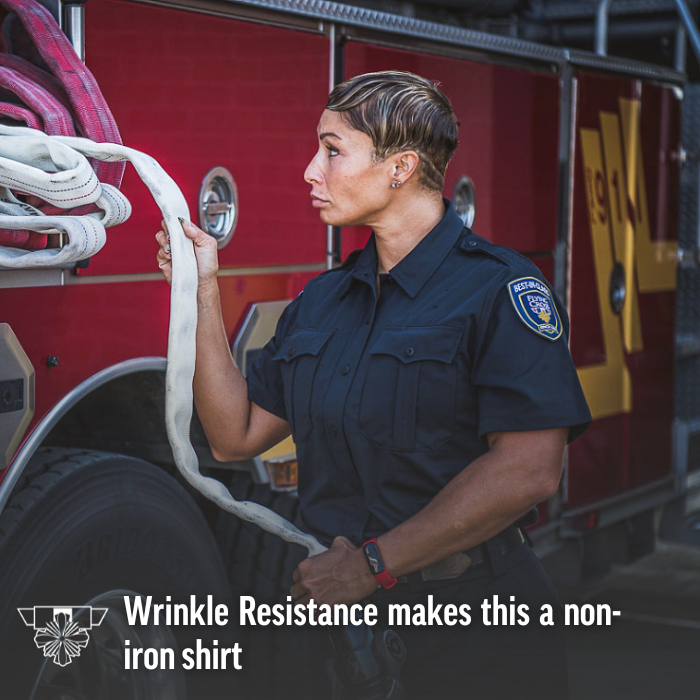 Photograph of a female firefighter in a wrinkle-resistant, dark blue uniform inspecting a fire hose next to a red firetruck with the number 1911 visible featuring a gold badge, a smartwatch and a white hose