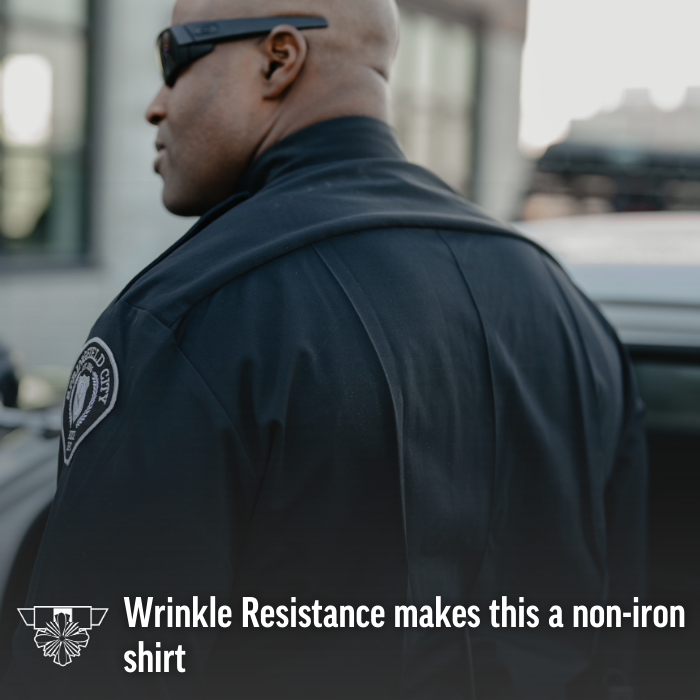 Photograph of a police officer wearing a wrinkle-resistant black uniform shirt with a city emblem featuring a shield and greenery, standing outdoors near a building and a vehicle, showcasing the fabric's smooth texture and dark color