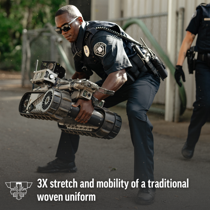 Photograph a police officer in a dark uniform carrying a tracked robot outdoors with gray pavement and blurred background buildings showcasing the device's dark gray metallic body, multiple cameras, and the officer's dark sunglasses and wristwatch.