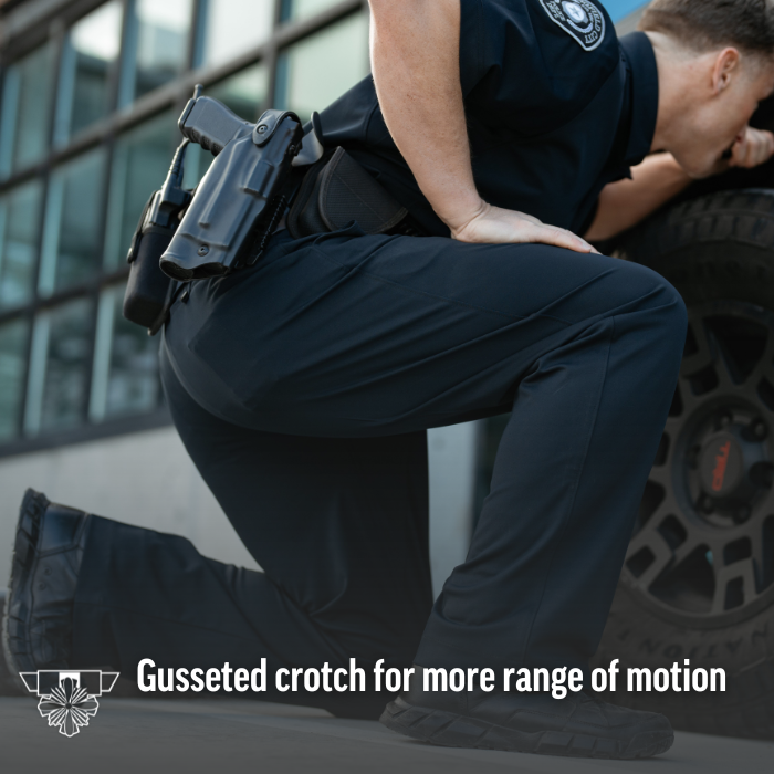 Photograph of a police officer in dark uniform kneeling near a vehicle next to a modern building with large glass windows showcasing a black firearm in a holster, tactical boots, and a gusseted crotch for increased mobility
