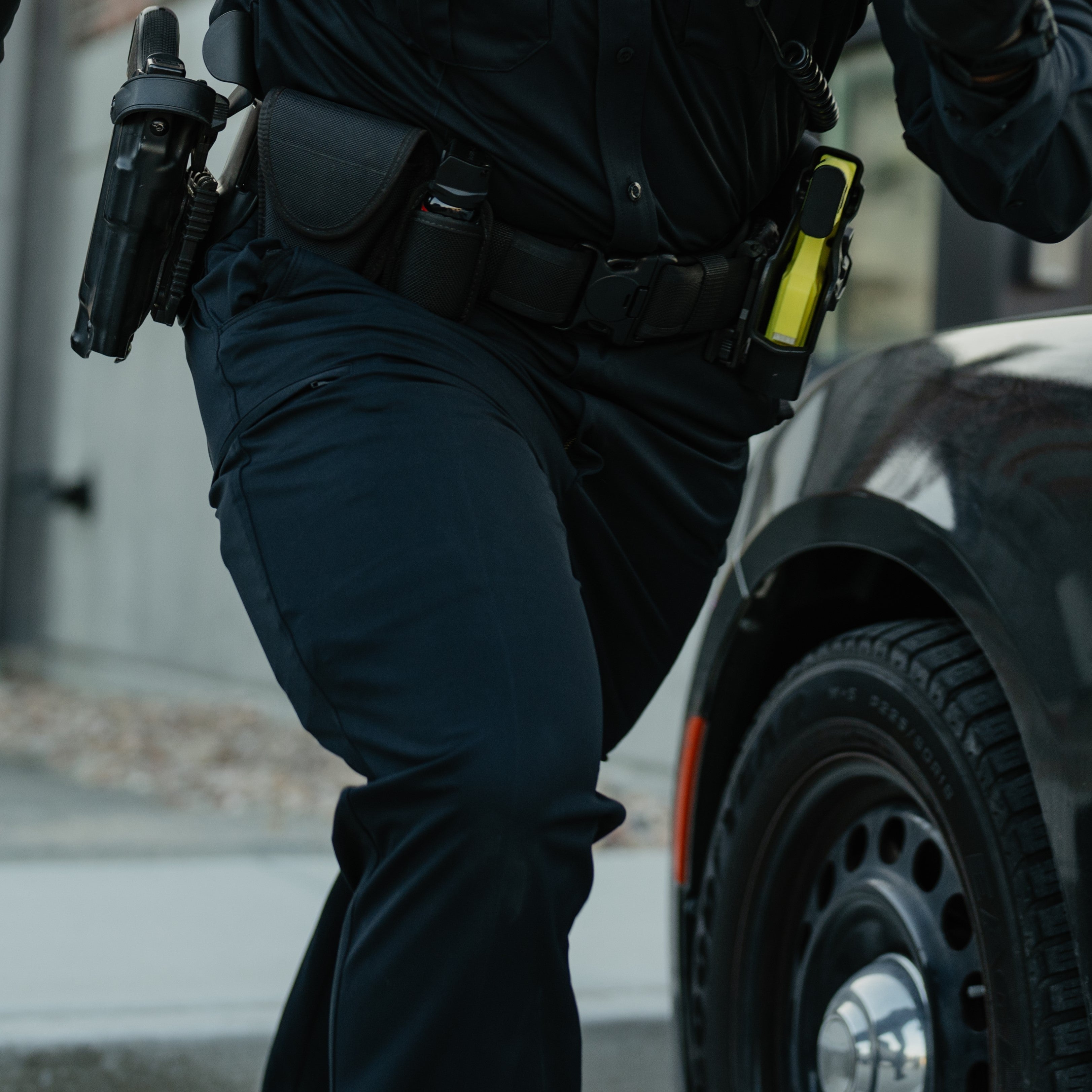 Photograph of a police officer in motion near a vehicle showing a black firearm in a holster a yellow taser and dark uniform pants