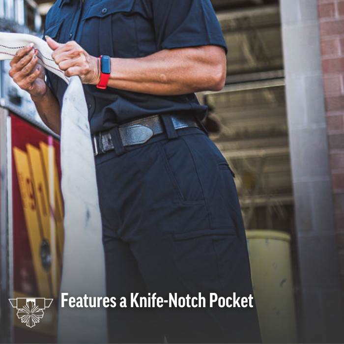 Photograph of a firefighter in dark uniform pants and a red smartwatch holding a firehose near a firetruck with red and yellow markings and a brick building showing a knife-notch pocket detail