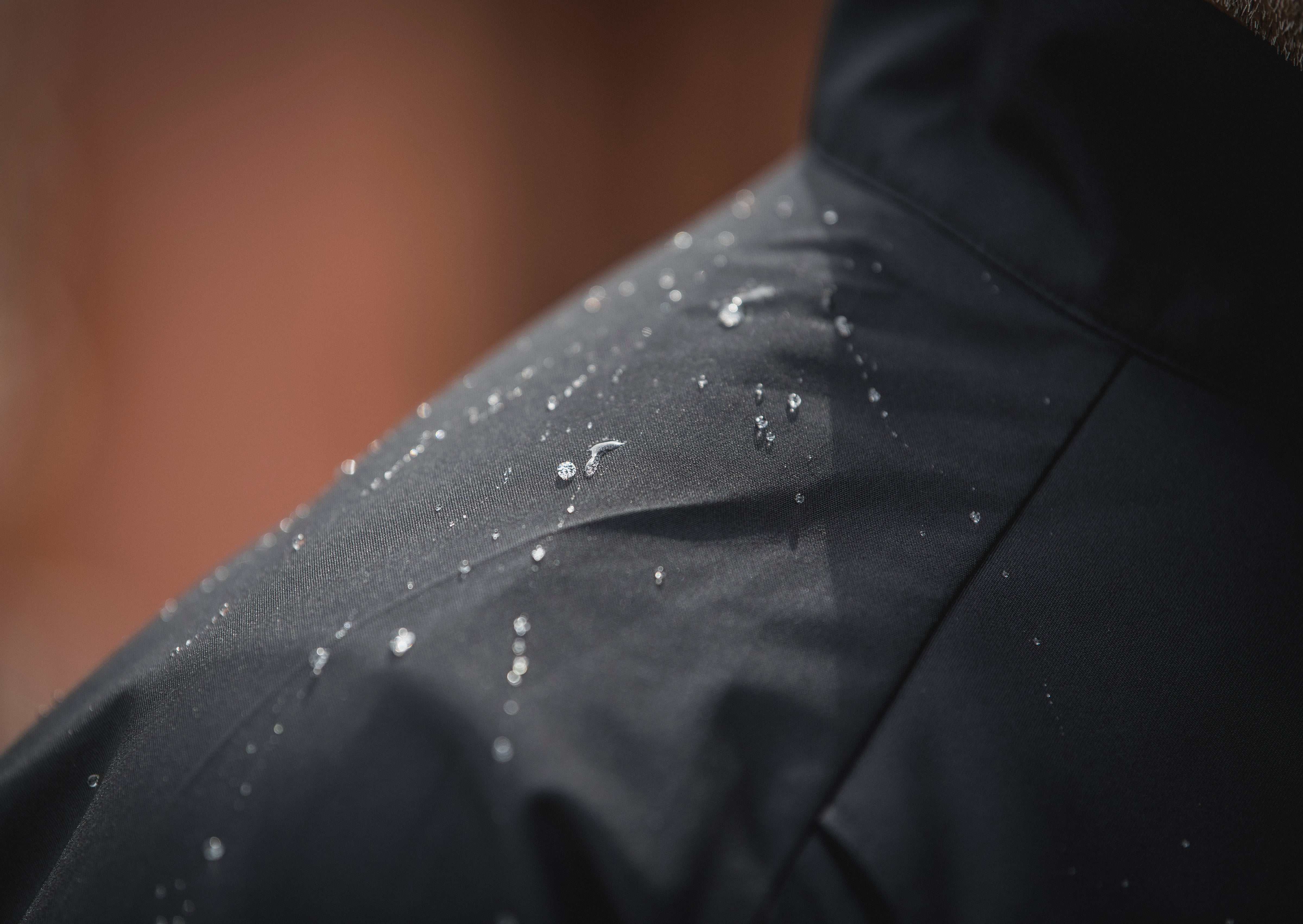 Close-up photograph showing water droplets on dark gray fabric with a blurred reddish-brown background, highlighting the fabric's water-resistant texture and the glistening, reflective quality of the droplets.