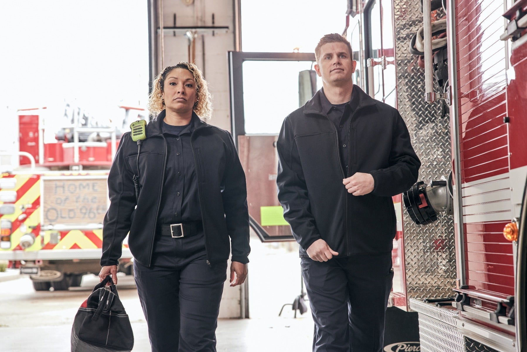 Photograph of two firefighters in a fire station, walking past a red firetruck with yellow and red chevron stripes and the words Home of Old 96, one firefighter carries a black bag and wears a lime green radio, showcasing their dark uniforms and serious expressions