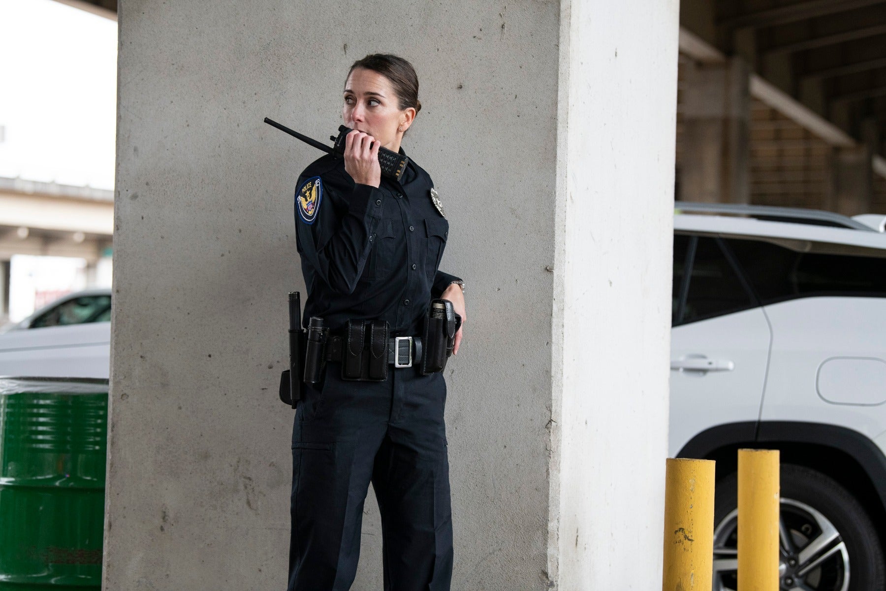 Photograph of a police officer using a radio in a parking garage near a white SUV and concrete pillars featuring a police badge a utility belt and a dark uniform