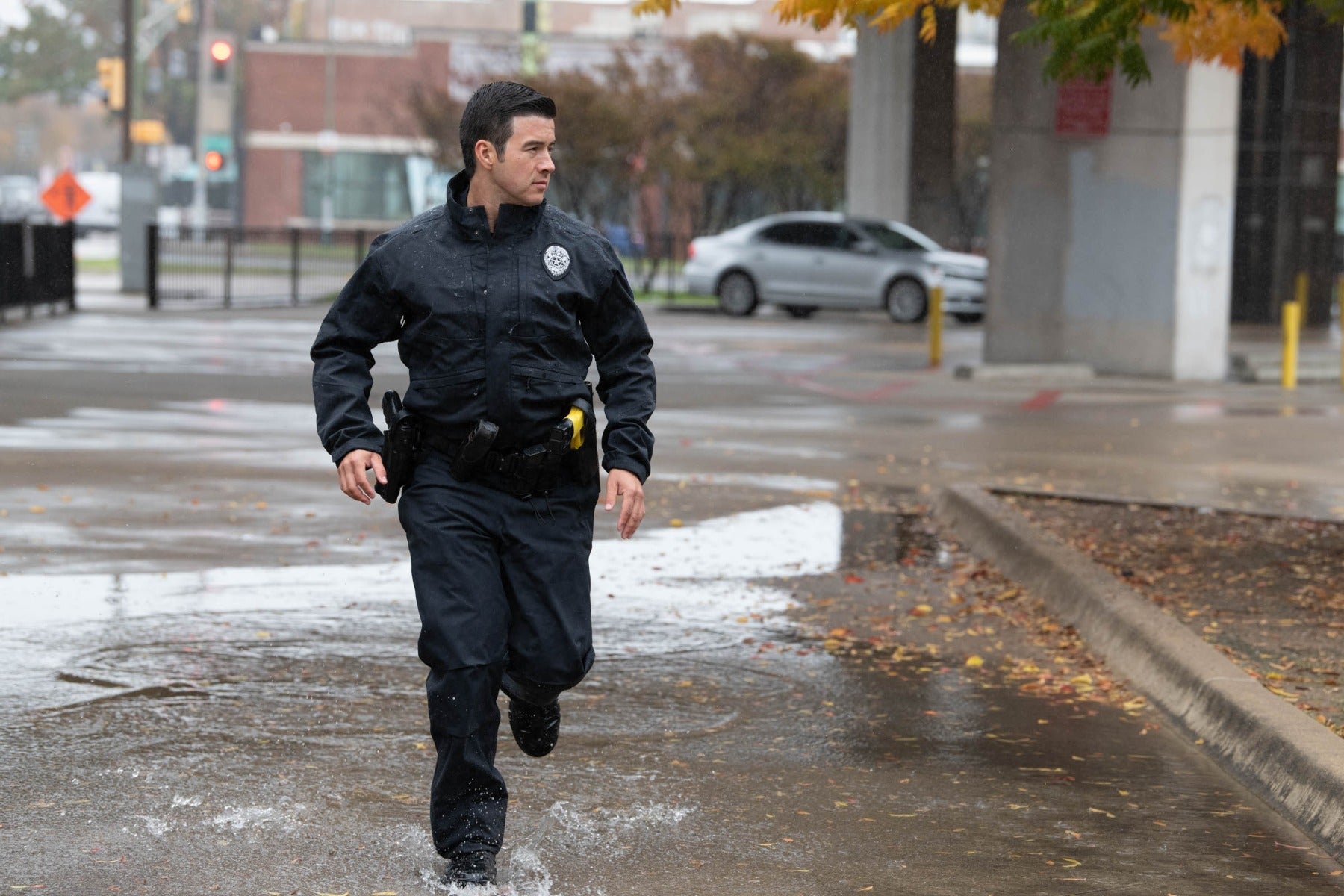 Photograph Still from TV series, a police officer runs through a rain-soaked city street near a traffic light and modern buildings with autumn leaves and dark puddles reflecting the overcast sky