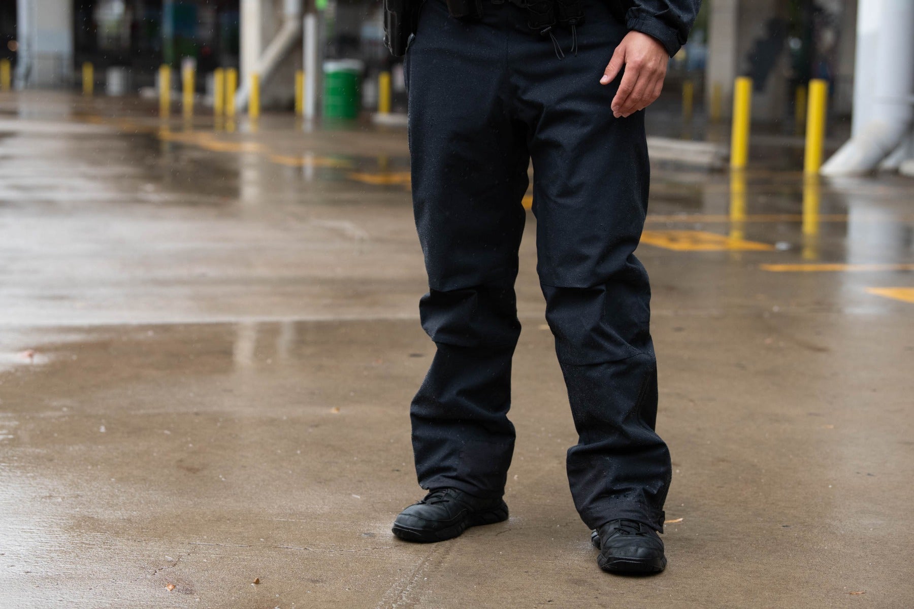 Photograph lower body of a person wearing black tactical pants and shoes in a rain-soaked parking lot with yellow parking dividers and a green trash can in the background