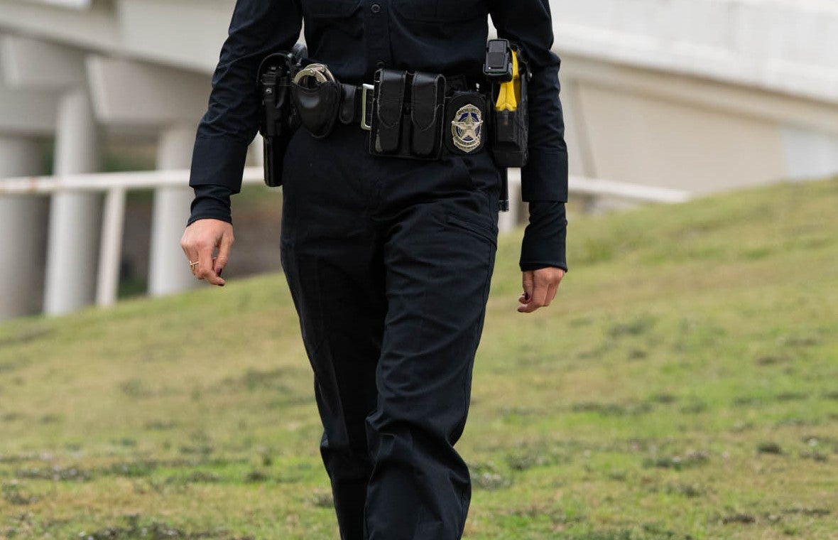 Photograph of a police officer in uniform walking on a grassy hill near a modern bridge with white columns and pillars a gold ring and a yellow utility device are visible on her belt