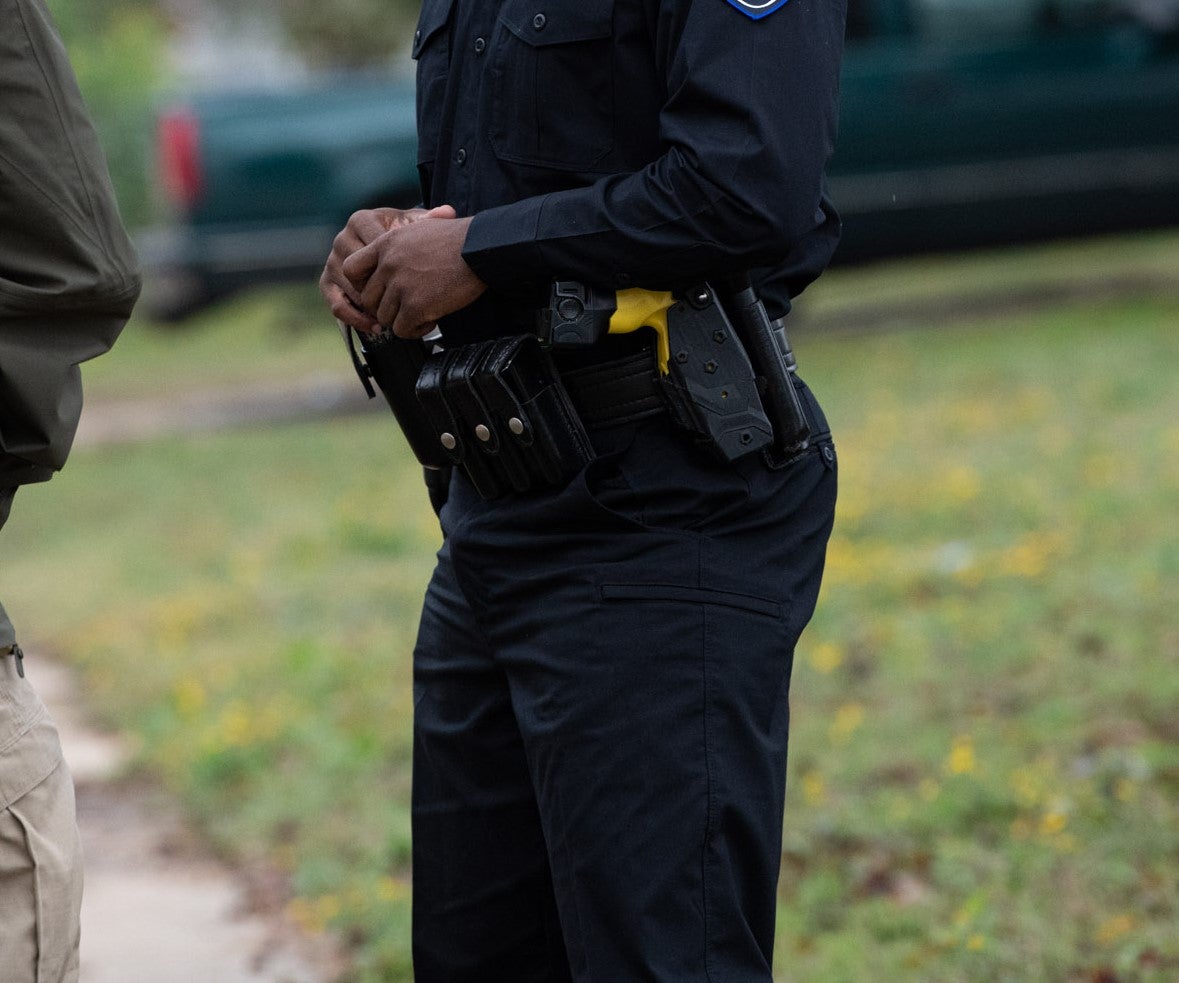 Photograph of a police officer in uniform outdoors with a handgun and multiple magazine pouches on their duty belt a blurred out green vehicle and grassy area are in the background