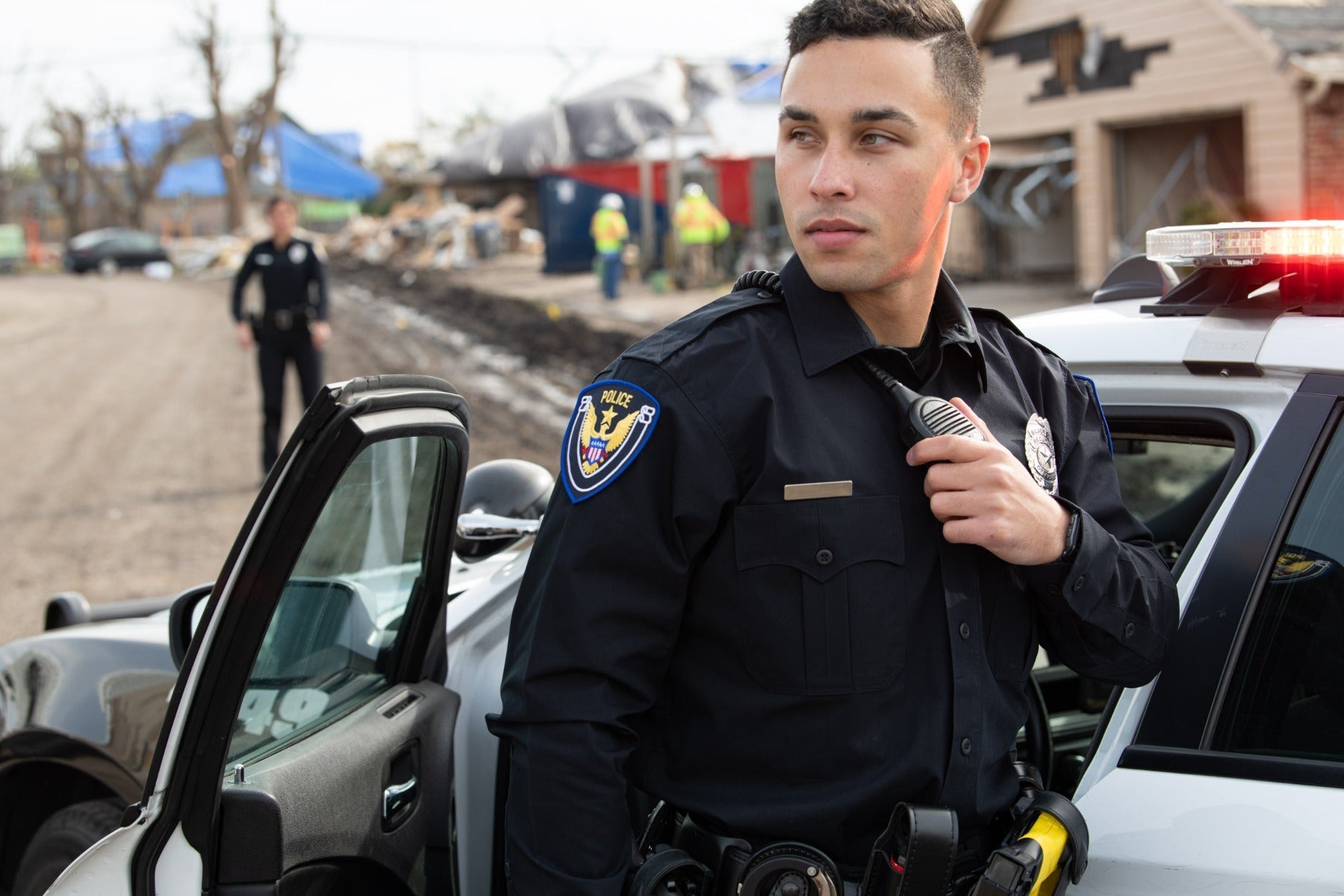 Photograph Police officer by unknown artist a male officer using a radio stands next to his police car amidst a disaster zone featuring damaged homes blue tarps and debris a police shield patch and a yellow utility tool are visible