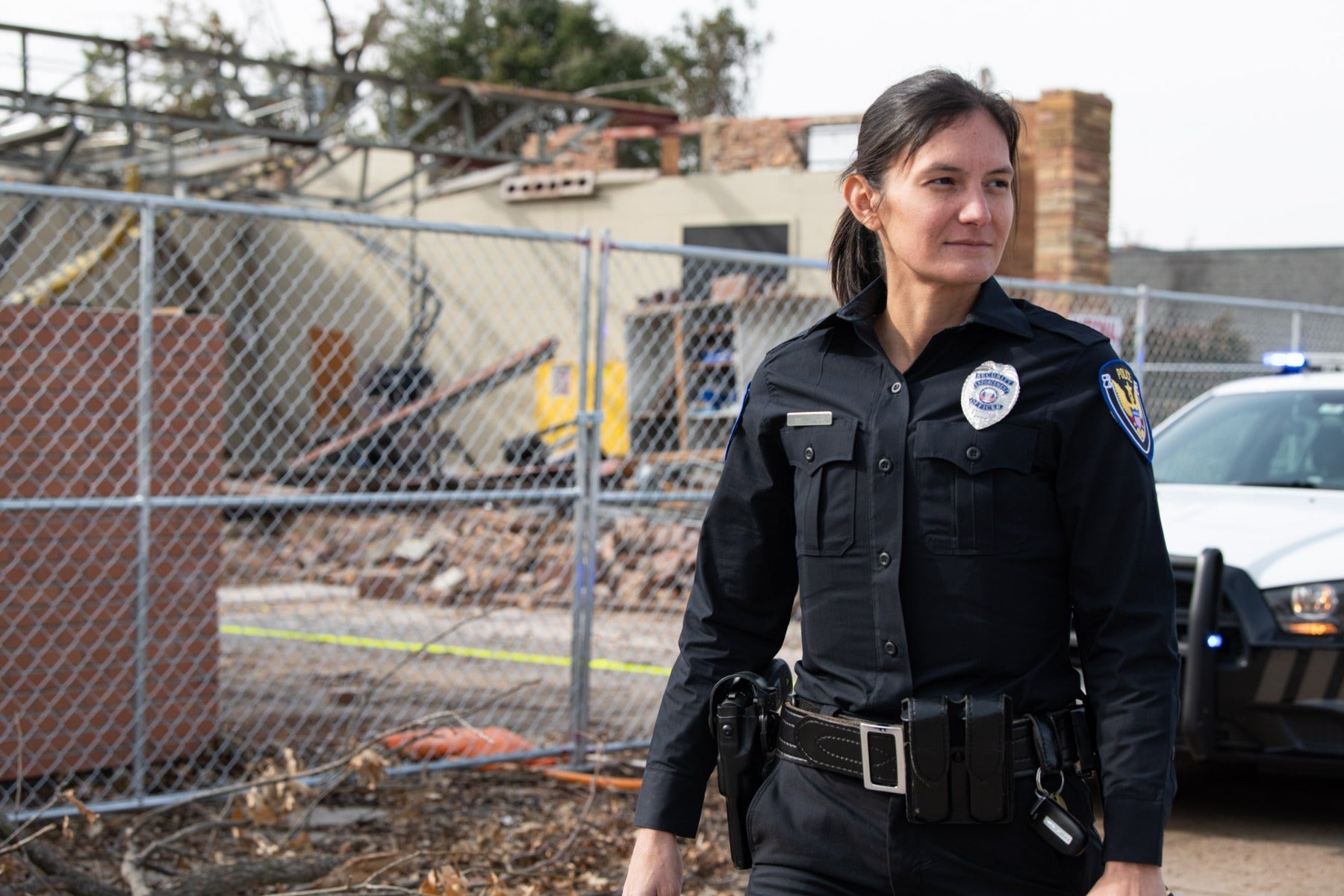 Photograph of a female police officer in uniform standing near a chain link fence surrounding a damaged building with exposed brick and a partially visible police car featuring a blue light and a silver badge with colorful detailing.