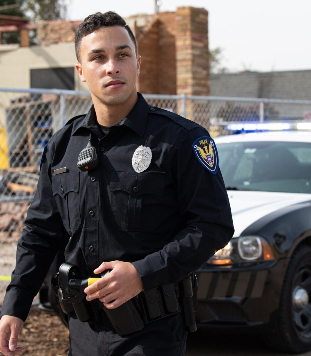 Photograph of a Silver Star County police officer in uniform near a damaged building and police car featuring a black uniform, a silver badge, and a yellow taser.