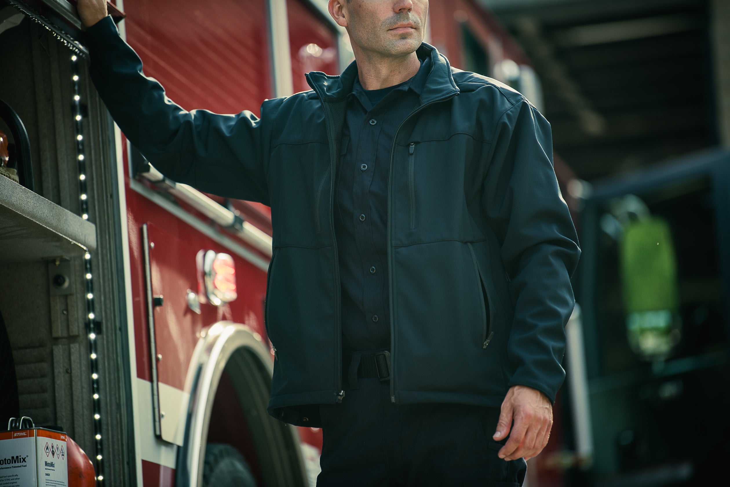 Photography: A firefighter in a dark jacket stands next to a red firetruck with a visible white stripe, LED lights, and a fuel can showcasing safety labels, his expression serious and his posture relaxed
