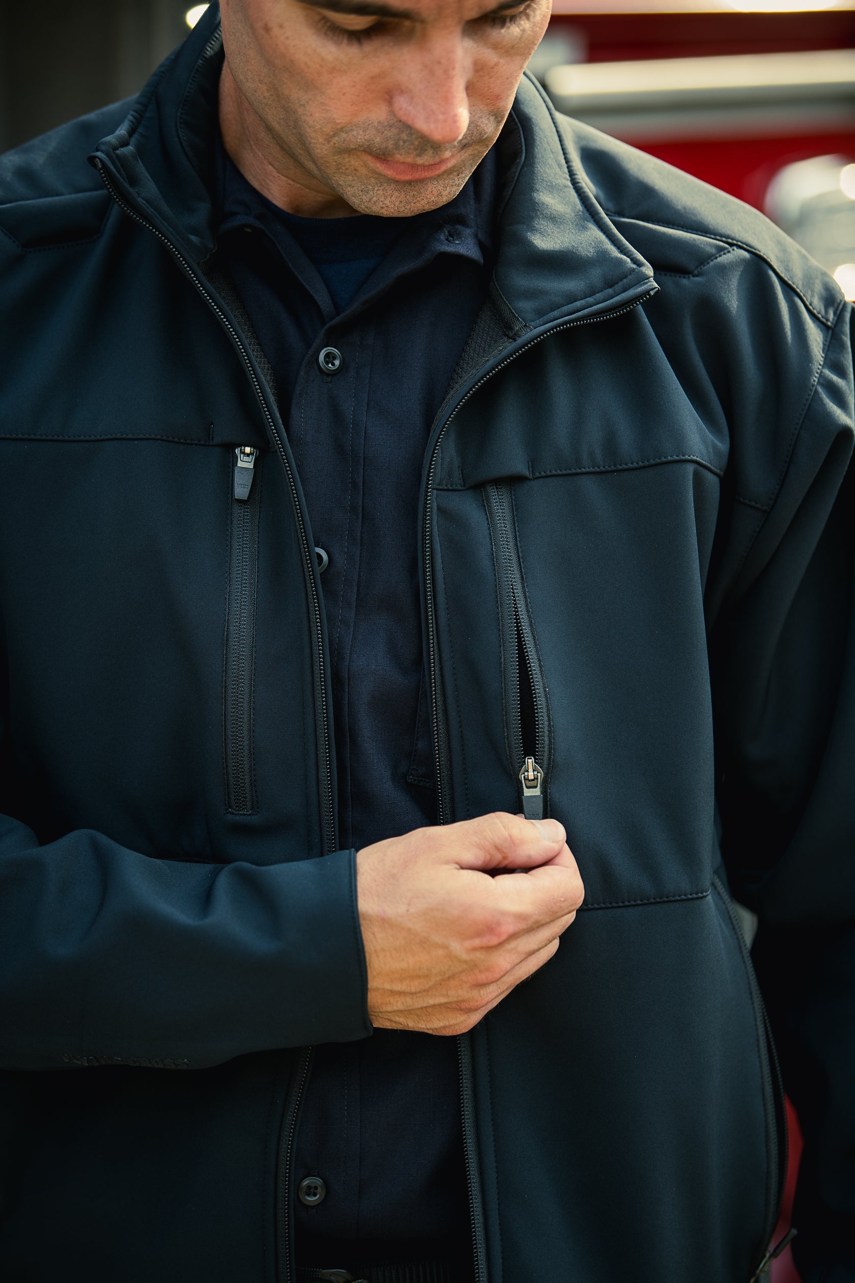 A photograph shows a man's dark navy soft-shell jacket with contrasting black zippers and stitching, a blurred red firetruck in the background