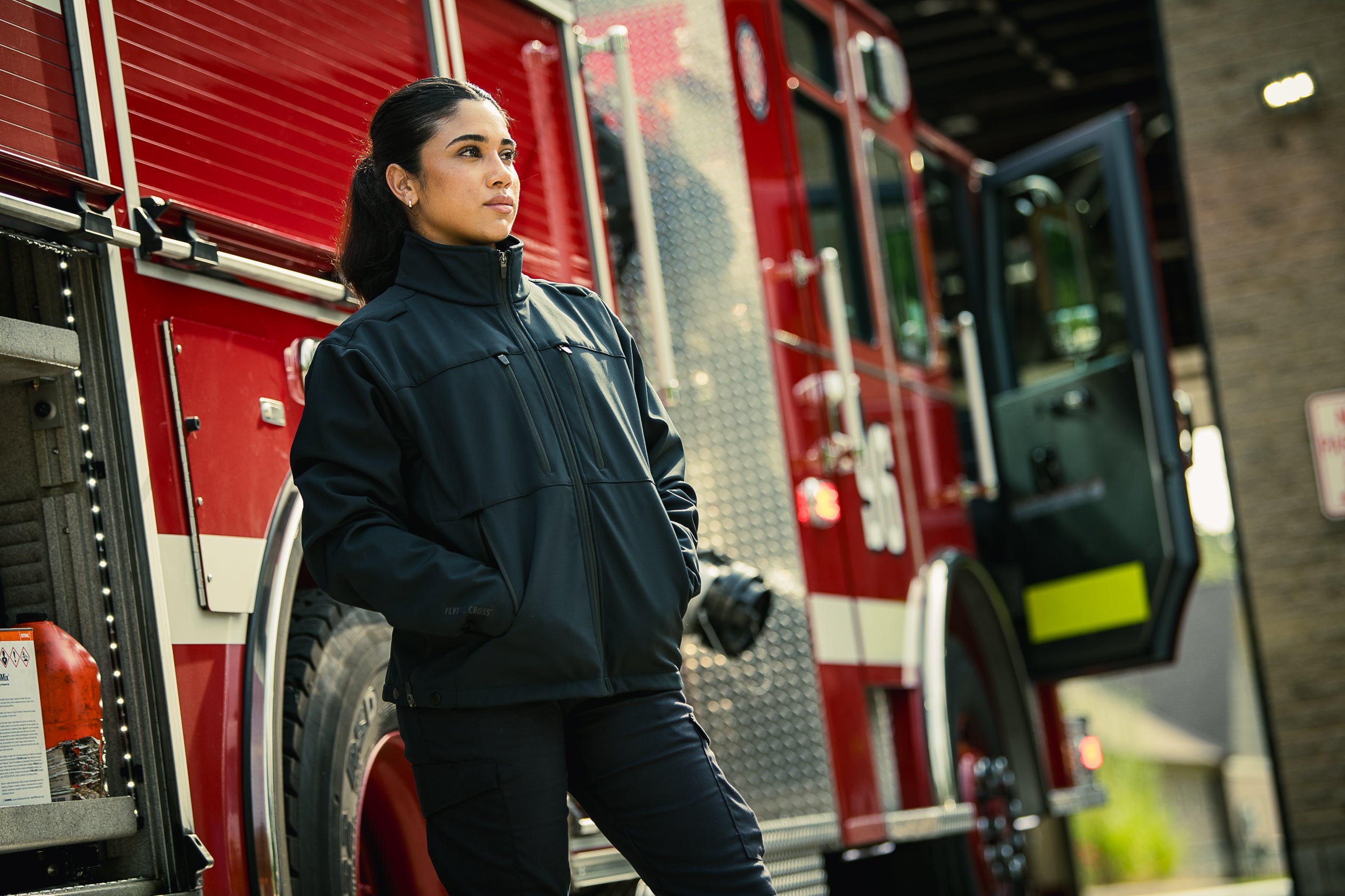 Photography unknown artist a female firefighter in a black jacket stands by a red firetruck at a fire station featuring white stripes and LED lights next to a partially visible hose and a blurred background