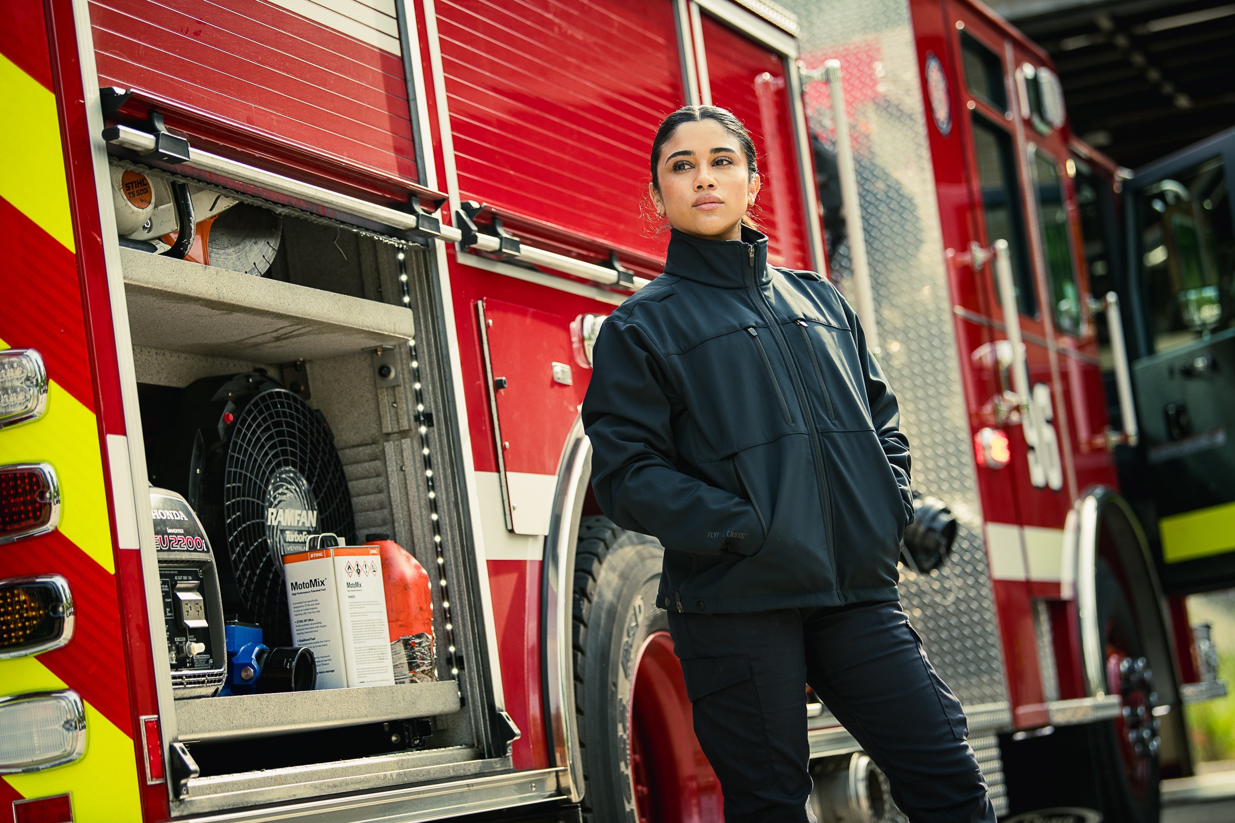 Photo of a woman in a black Fly Cross jacket standing by a red firetruck with visible equipment including a Stihl saw, Ramfan ventilator, and Honda generator, showcasing vibrant yellow and red details