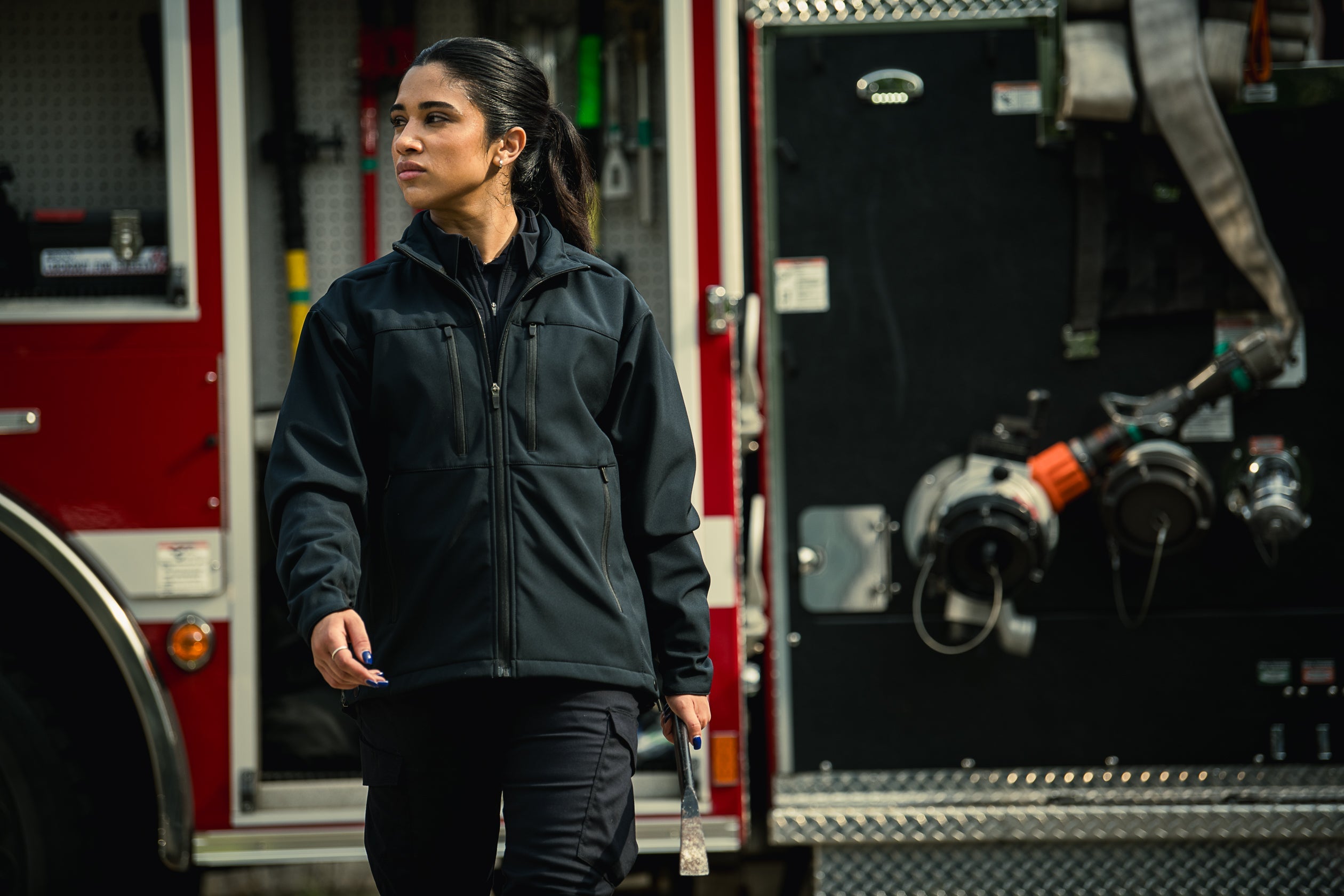 Photograph of a female firefighter in a black jacket and dark pants holding a halligan bar in front of a red firetruck with visible hoses and a diamond-plate surface, showcasing her serious expression, dark hair tied back and navy blue nail polish