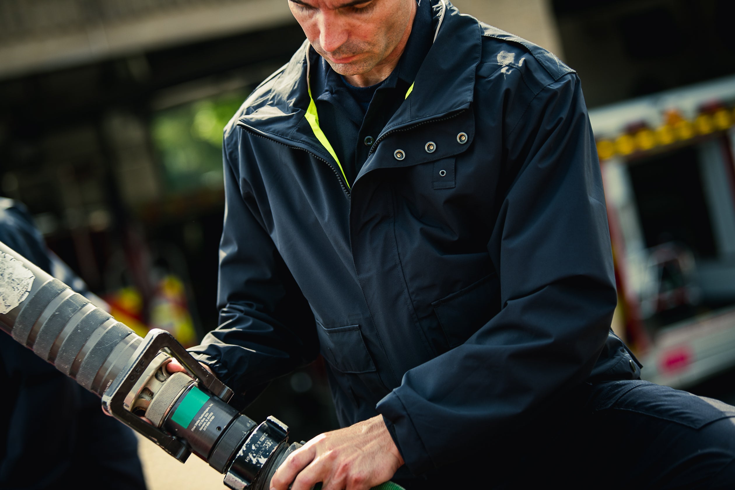 A photograph shows a firefighter in a dark navy jacket with a neon yellow inner lining connecting a firehose nozzle near a firetruck with visible warning lights and reflectors