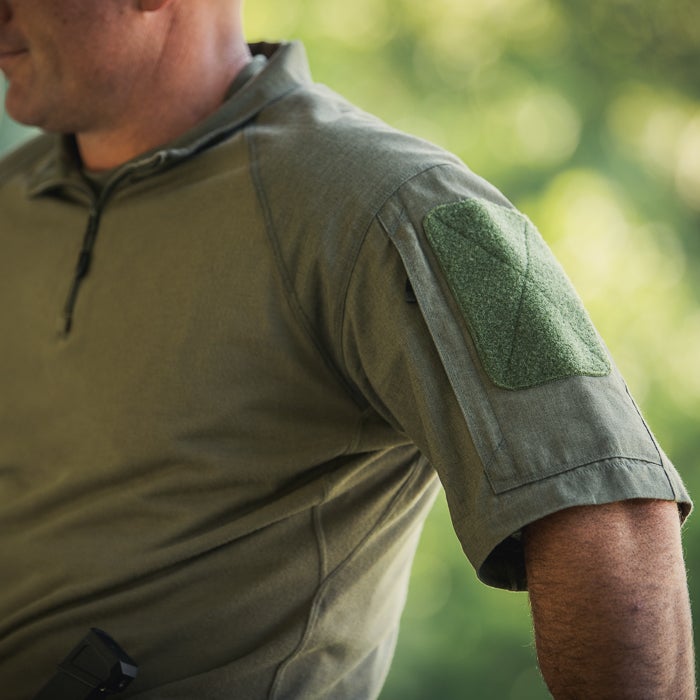 Photograph showing a man's olive green tactical shirt with a dark green Velcro patch on the sleeve and a hint of a firearm in the pocket against a blurred natural background