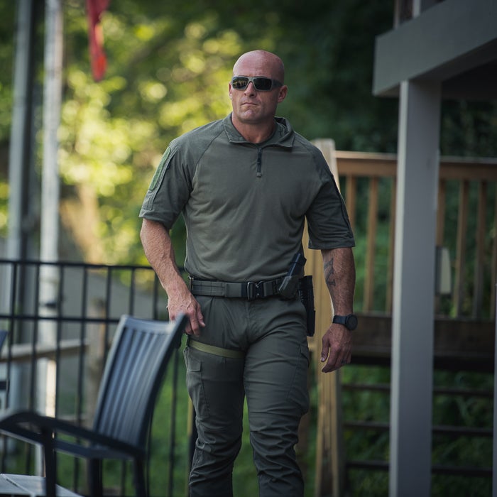Photograph of a man in a green tactical shirt and pants walking near a house and fence wearing sunglasses and a watch