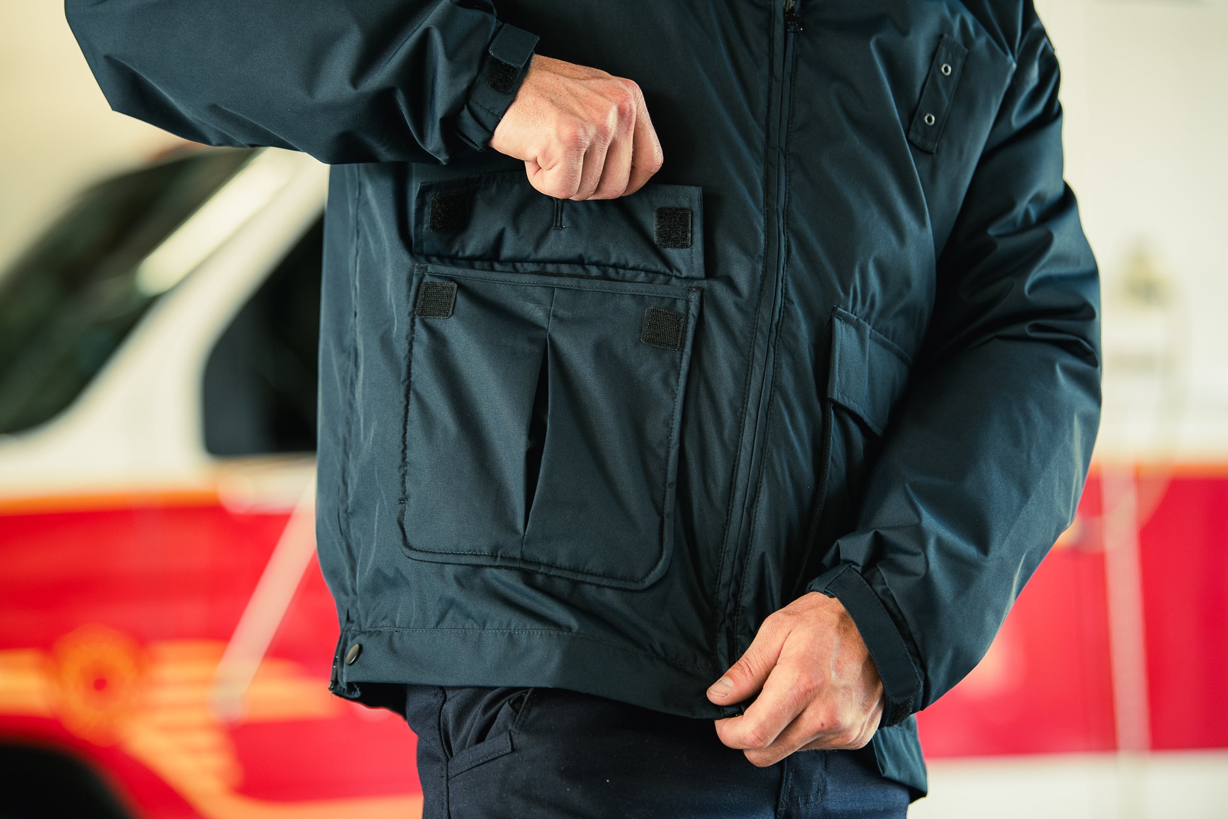 Photograph showing a dark navy blue jacket with multiple pockets and Velcro closures a blurred red and white ambulance is in the background