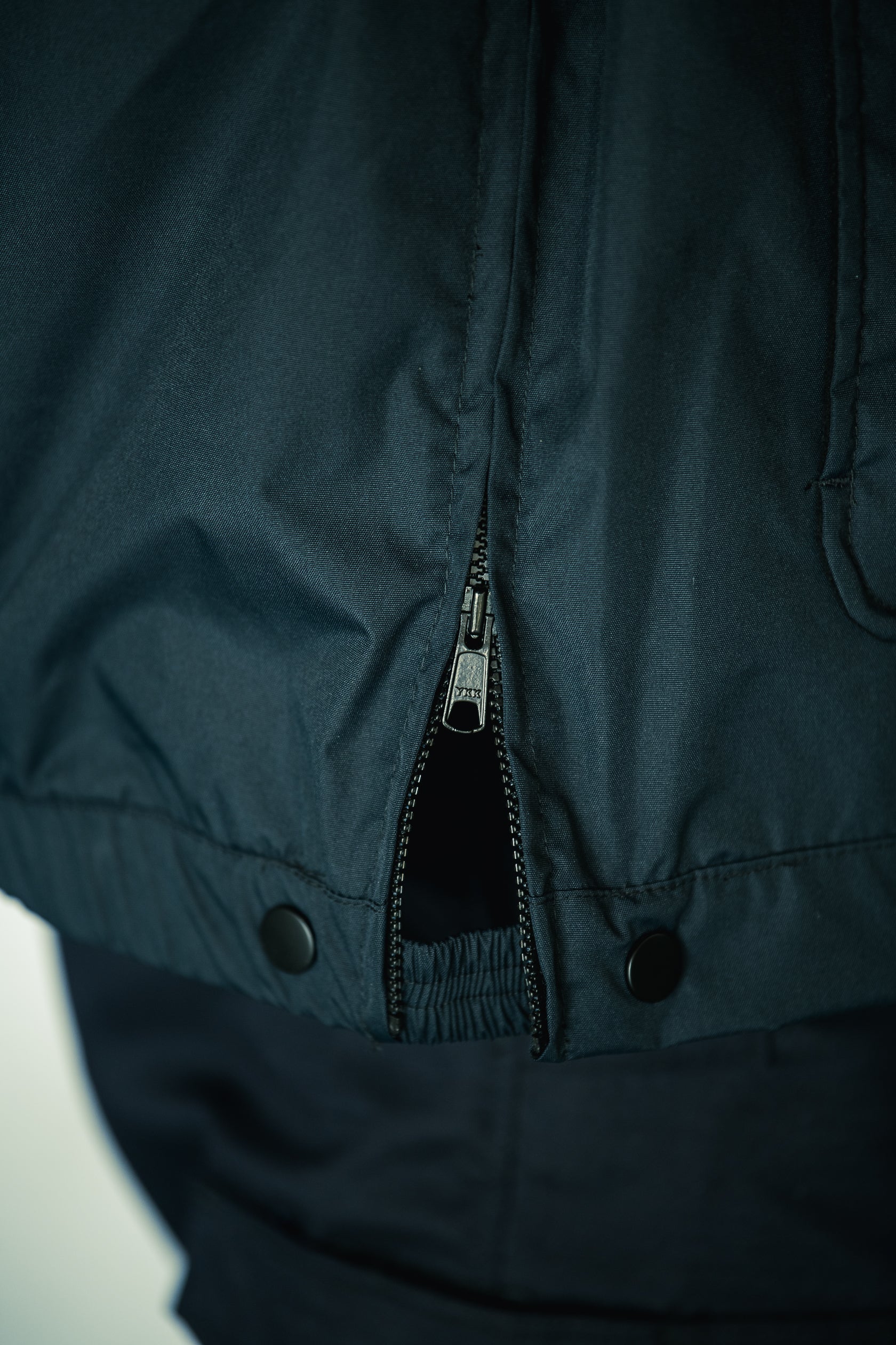 Close-up photograph showing the dark teal fabric texture of a jacket's zipper and snap buttons detail a YKK zipper pull and ribbed waistband