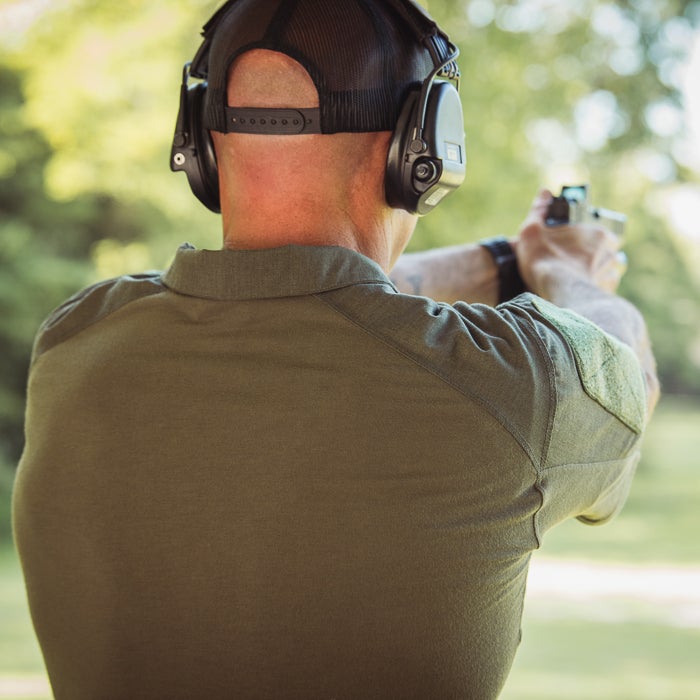 Photograph Man wearing olive green shirt and black ear protection shooting a handgun at an outdoor range with blurred green background