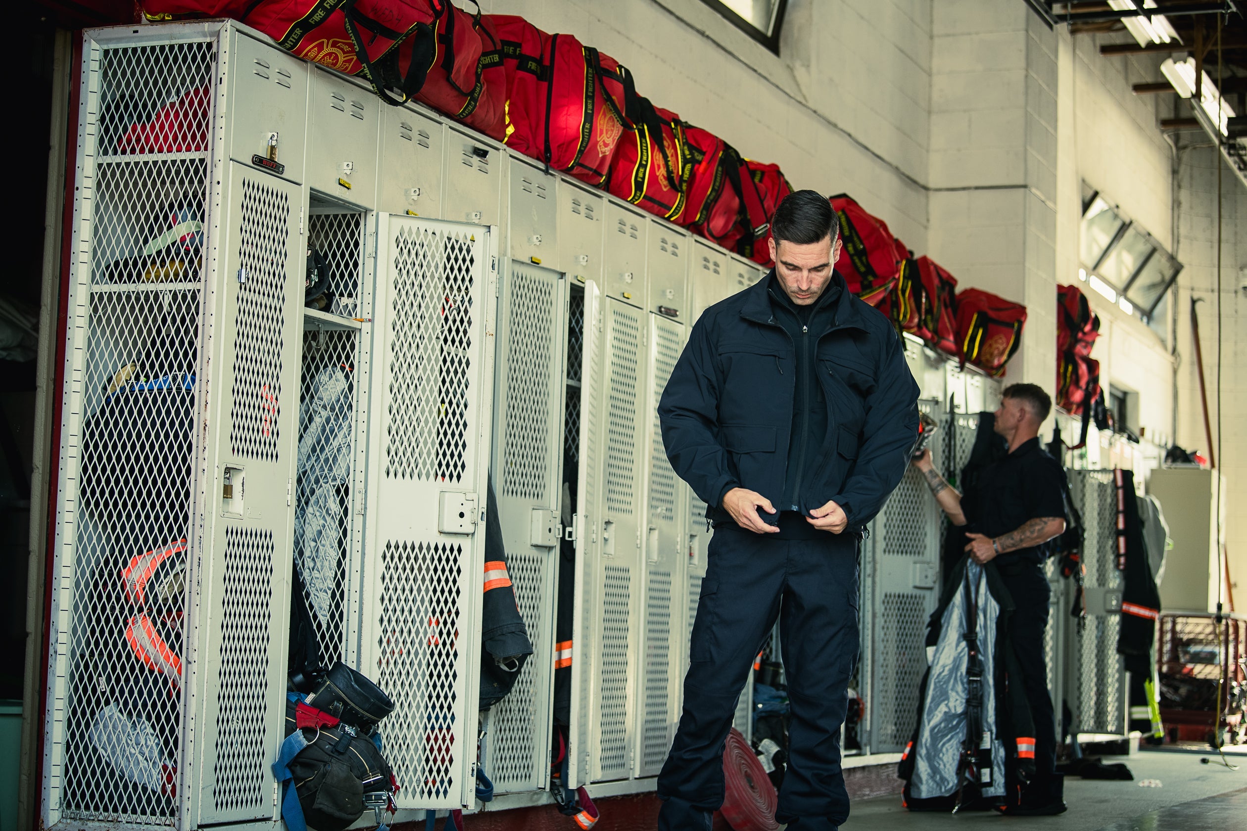 Photograph of a firefighter in a dark jacket and pants in a fire station locker room, amidst red fire bags, gray metal lockers, and a partially visible colleague with tattoos.